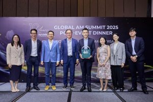 A group of people standing together indoors for a photo. One person in the center is holding a 3D-printed tactile braille storybook. The setting includes a backdrop with signage indicating NAMIC Singapore’s 10th Anniversary Dinner.