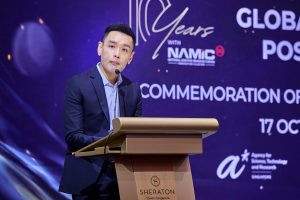 A person in a dark suit standing at a podium with a microphone, giving a speech. The podium has the Sheraton Towers Singapore logo. Behind is a purple backdrop with text that reads ‘10 Years with NAMIC,’ ‘Global,’ and ‘Commemoration of,’ along with the date 17 October and the Agency for Science, Technology and Research logo.