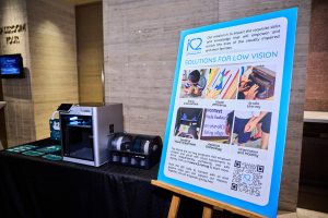 A poster titled ‘Solutions for Low Vision’ displayed on an easel next to a 3D printer on a black table. The poster includes images and text about tactile interaction, visual efficiency, braille literacy, and assistive technology.