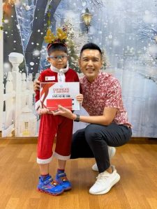 A child dressed in a festive red Santa outfit with reindeer antlers stands next to an adult in a red patterned shirt. They are holding a Certificate of Achievement and gift vouchers during a sports awards presentation. The background features a winter-themed backdrop with snow-covered scenery.