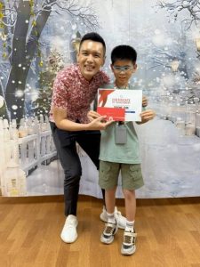 A child stands next to an adult while holding a Certificate of Achievement during a sports awards presentation. Both are positioned in front of a winter-themed backdrop featuring snow-covered scenery and decorative lights. The child is wearing a green T-shirt and shorts with sneakers, and the adult is dressed in a red patterned shirt and dark pants.