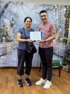 A volunteer stands next to an adult while holding a Certificate of Appreciation during a presentation. Both are positioned in front of a winter-themed backdrop featuring snow-covered trees, decorative lights, and a Christmas tree partially visible on the right.