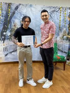 A volunteer stands next to an adult while holding a Certificate of Appreciation during a presentation. Both are positioned in front of a winter-themed backdrop featuring snow-covered trees, decorative lights, and a Christmas tree partially visible on the right.