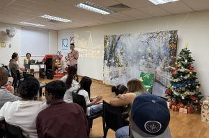 A person standing at the front of a room giving a welcome speech during a Christmas party. The room is decorated with a Christmas tree, wrapped gifts, and a winter-themed backdrop. Several people are seated and facing the speaker.