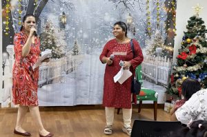 Two individuals standing at the front of a room acting as emcees during a Christmas party. They are dressed in festive red outfits and holding microphones and papers. Behind them is a winter-themed backdrop and a decorated Christmas tree with wrapped gifts. Several chairs and attendees are visible in the foreground.