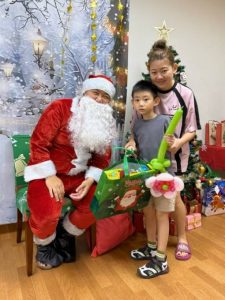 Santa Claus poses with a child beneficiary and their family during a Christmas celebration. The group stands in front of a winter-themed backdrop with a decorated Christmas tree and wrapped gifts, creating a festive atmosphere.
