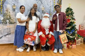 Santa Claus poses with a child beneficiary and their family during a Christmas celebration. The group stands in front of a winter-themed backdrop with a decorated Christmas tree and wrapped gifts, creating a festive atmosphere.