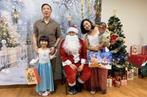 Santa Claus poses with a child beneficiary and their family during a Christmas celebration. The group stands in front of a winter-themed backdrop with a decorated Christmas tree and wrapped gifts, creating a festive atmosphere.