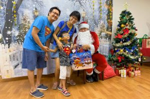 Santa Claus poses with a child beneficiary and their family during a Christmas celebration. The group stands in front of a winter-themed backdrop with a decorated Christmas tree and wrapped gifts, creating a festive atmosphere.
