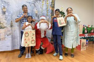 Santa Claus poses with a child beneficiary and their family during a Christmas celebration. The group stands in front of a winter-themed backdrop with a decorated Christmas tree and wrapped gifts, creating a festive atmosphere.