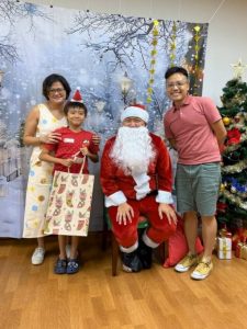 Santa Claus poses with a child beneficiary and their family during a Christmas celebration. The group stands in front of a winter-themed backdrop with a decorated Christmas tree and wrapped gifts, creating a festive atmosphere.