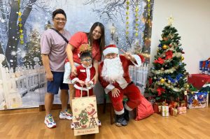 Santa Claus poses with a child beneficiary and their family during a Christmas celebration. The group stands in front of a winter-themed backdrop with a decorated Christmas tree and wrapped gifts, creating a festive atmosphere.