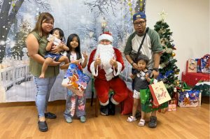 Santa Claus poses with a child beneficiary and their family during a Christmas celebration. The group stands in front of a winter-themed backdrop with a decorated Christmas tree and wrapped gifts, creating a festive atmosphere.