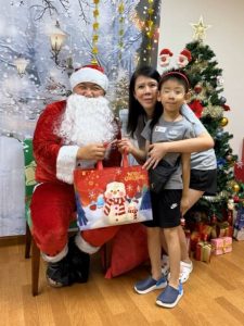 Santa Claus poses with a child beneficiary and their family during a Christmas celebration. The group stands in front of a winter-themed backdrop with a decorated Christmas tree and wrapped gifts, creating a festive atmosphere.