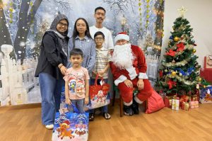 Santa Claus poses with a child beneficiary and their family during a Christmas celebration. The group stands in front of a winter-themed backdrop with a decorated Christmas tree and wrapped gifts, creating a festive atmosphere.
