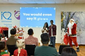 Two siblings stand at the front of a room singing Christmas songs into microphones during a festive event. A large screen behind them displays the lyrics ‘You would even say it glows.’ To the right, a person dressed as Santa Claus gestures cheerfully, holding a red sack. The setting includes a decorated Christmas tree, wrapped gifts, and a winter-themed backdrop. Audience members are seated in the foreground.