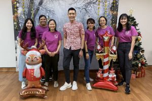 A group of seven people posing for a photo in front of a winter-themed backdrop during a Christmas celebration. Six individuals are wearing matching purple shirts with the iC2 logo, and one person in the center is dressed in a red patterned shirt. Festive decorations include a Christmas tree with ornaments and presents, as well as two large inflatable figures—a reindeer and a candy cane lamp post.