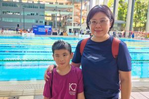 A child wearing a purple iC2 T-shirt stands beside an adult near an outdoor swimming pool at a sports venue. The pool has multiple lanes with green and yellow lane dividers, and a competition banner is visible in the background. The adult has a backpack, and the setting includes tall columns, buildings, and trees under bright daylight.