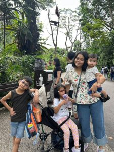 A family group at the zoo, standing on a pathway surrounded by lush greenery. One child is leaning on a stroller with a drink bottle, another is seated in the stroller holding a snack, and an adult is carrying a toddler.