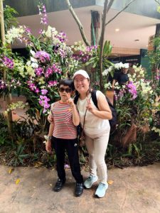Two individuals posing in front of a vibrant display of orchids at the zoo. The flowers include shades of purple, white, and pink, arranged among green foliage.