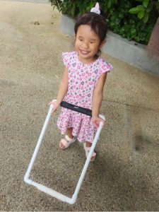 A young child wearing a pink leopard-print dress and sandals, holding a white rectangular frame with a black strap while standing on a textured pathway. Green foliage and a low concrete border are visible in the background.